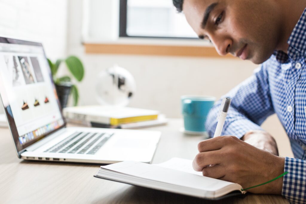 Como investir em Fiis: Vemos um homem de camisa quadriculada segurando uma caneta e anotando em um caderno em frente a um laptop.