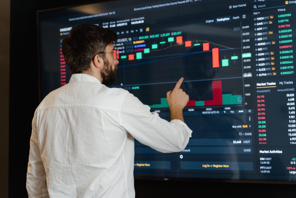 Home broker: vemos um homem de costa vestindo uma camisa branca e apontando para uma TV que exibe uma interface de home broker.