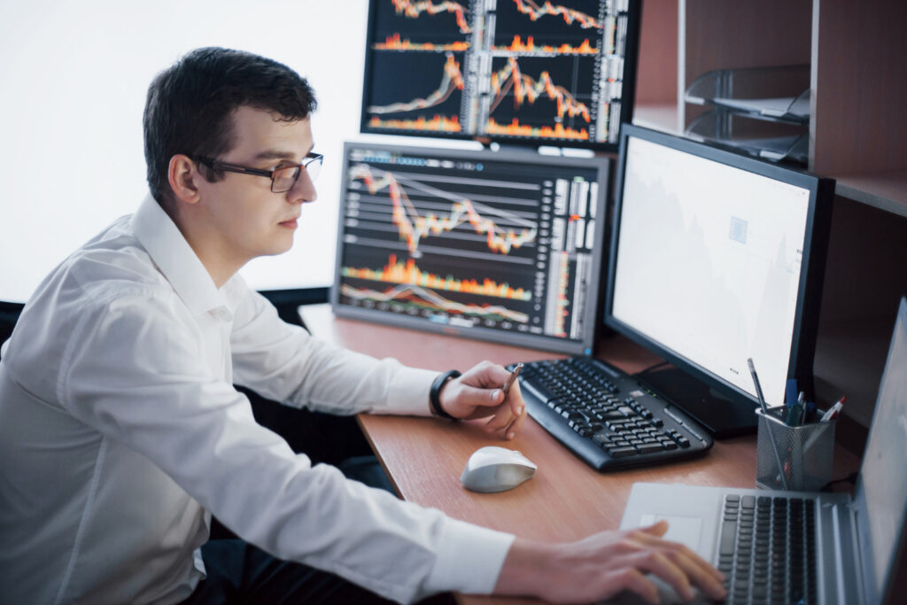 Homem está sentado em frente a um computador e tem mais dois monitores ao lado. Nas telas, aparecem gráficos da renda variável e o home broker que ele usa para negociar ativos