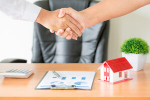 Dois executivos apertam as mãos sobre uma prancheta com papéis e uma miniatura de casa sinalizando o investimento em CRI