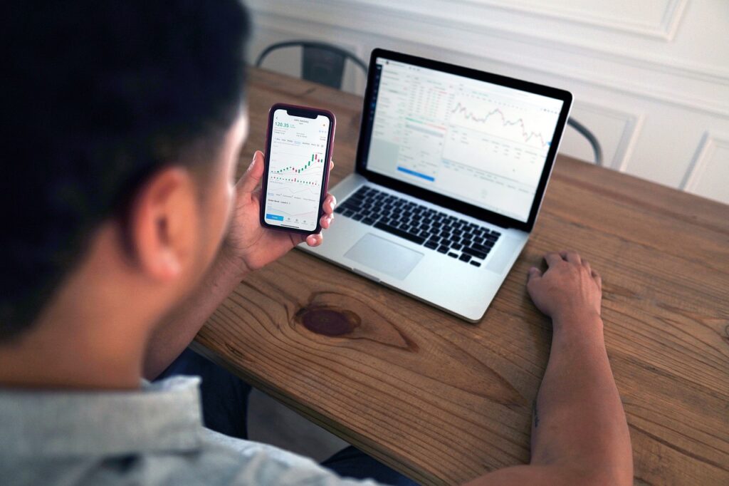 Homem segura o celular sentado em uma mesa de frente para o notebook, em ambas as telas aparecem gráficos de day trade