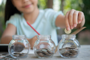 Menina asiática segura um lápis e coloca moedas em um dos três potinhos que aparece em sua frente para mostrar a diversificação possível com o perfil de investidor