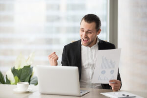 Executivo sentado à mesa em frente ao notebook segura uma folha de papel e comemora os ganhos de acordo com seu perfil de investidor